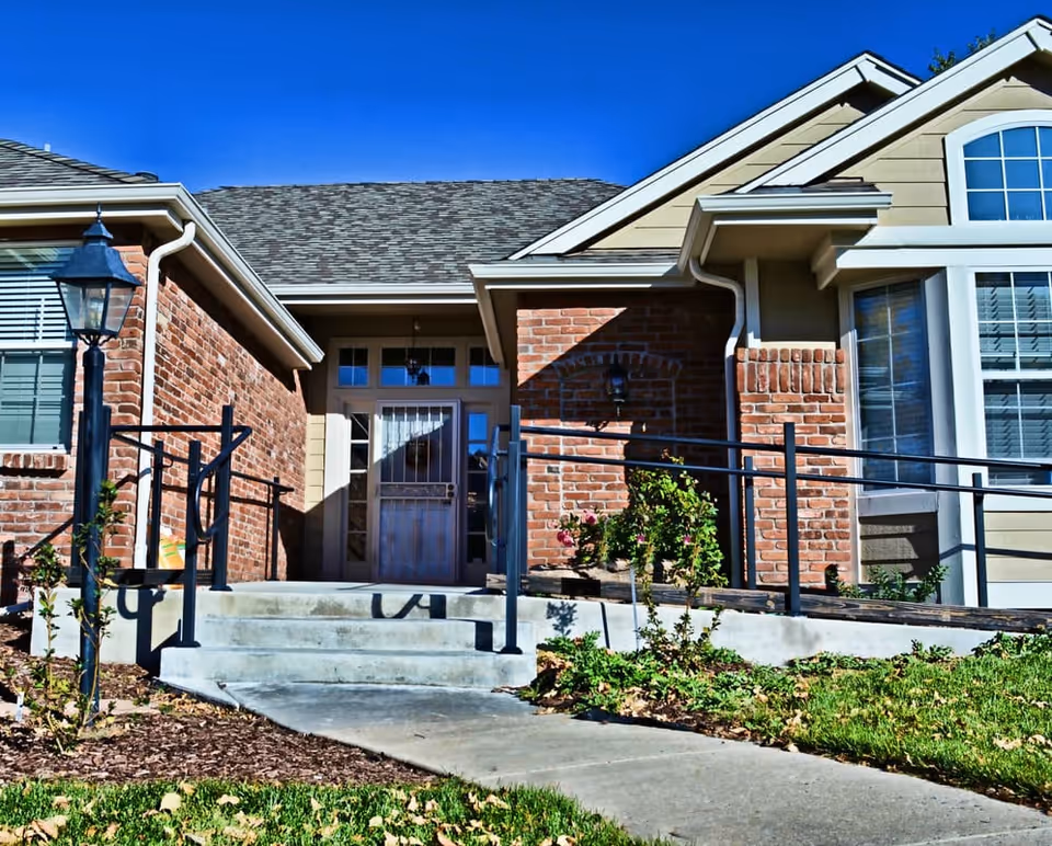 Front entrance of a brick and beige siding building with a concrete ramp and stairs leading up to a door with glass panels. There is a black metal handrail along the ramp and stairs, a lamp post on the left side, and some greenery and plants around the entrance under a clear blue sky.