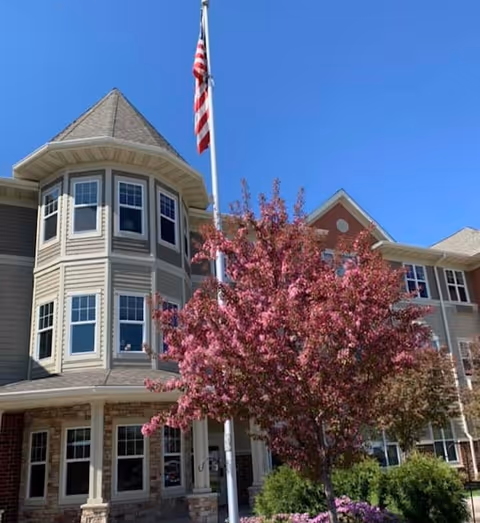 Exterior view of a multi-story senior living facility building with beige siding and stone accents. The building features a turret with multiple windows and a peaked roof. In front of the building, there is a flagpole with an American flag and a blooming pink tree surrounded by green shrubs under a clear blue sky.