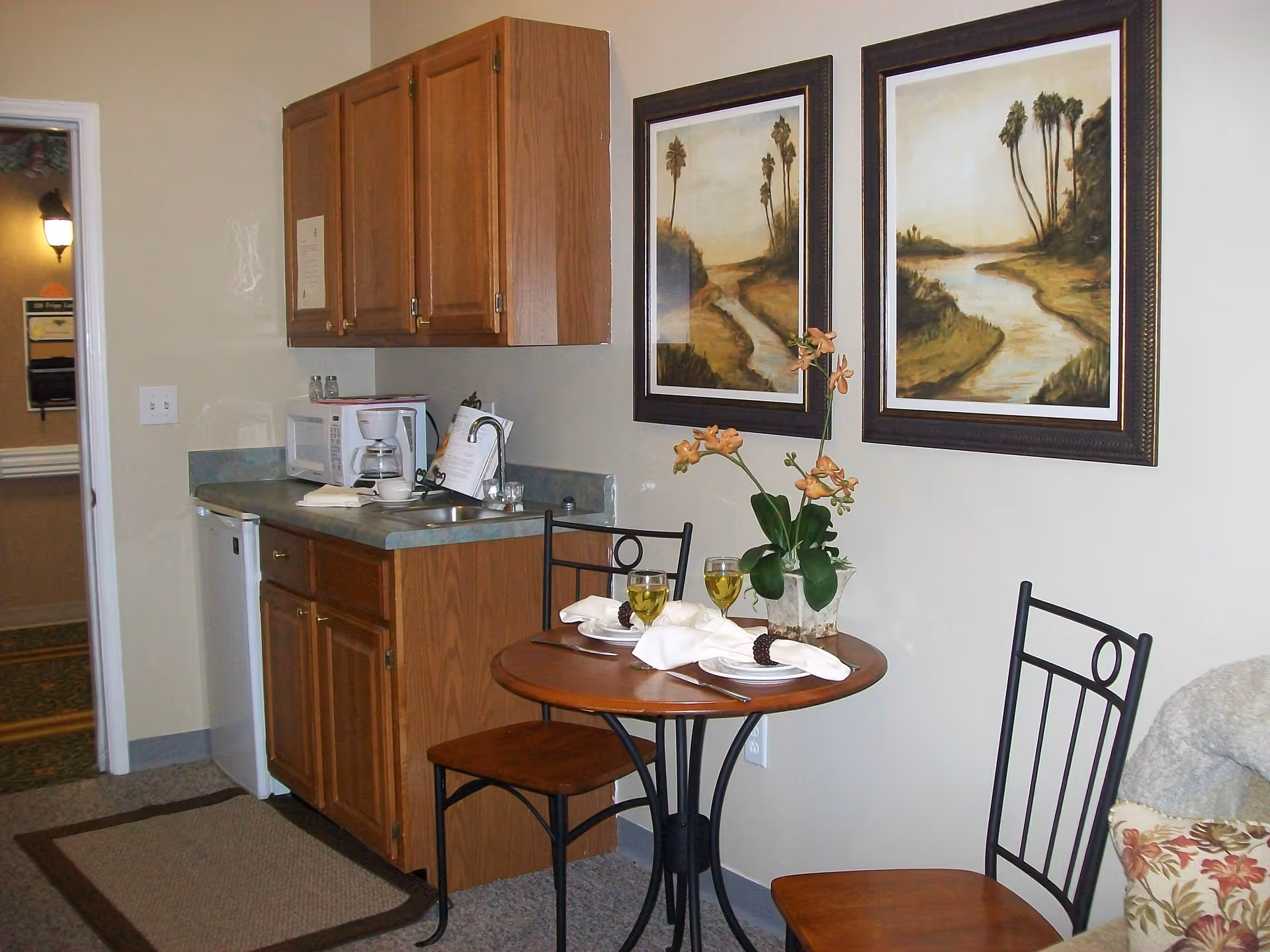 Small kitchenette and dining nook with a round table set for two, wooden cabinets, and framed landscape paintings on the wall.