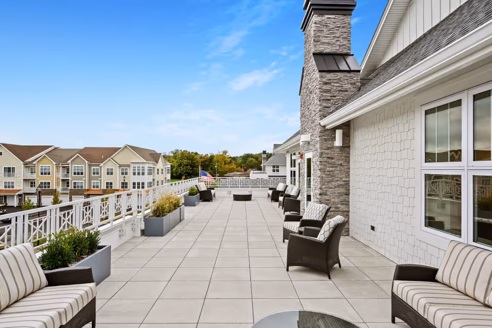 Spacious outdoor patio area with cushioned wicker chairs and sofas arranged along the sides. The patio has large square tiles and is decorated with rectangular planters containing greenery. The building exterior features stone and shingle siding with large windows. In the background, there are residential buildings and a clear blue sky.