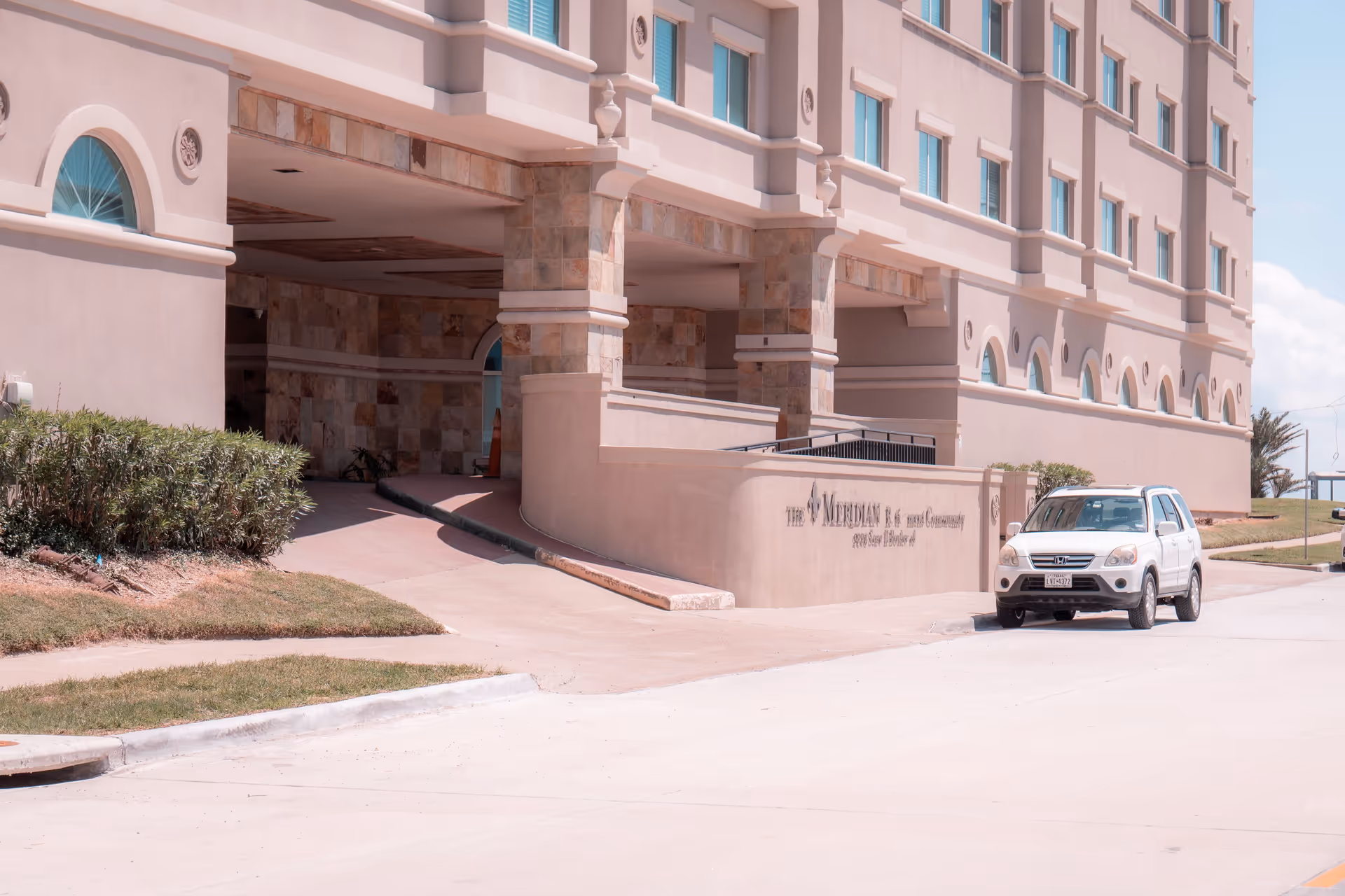 Front entrance of The Meridian senior living building showing a covered drop-off with a ramp, signage on a stucco wall, and a white SUV parked nearby.