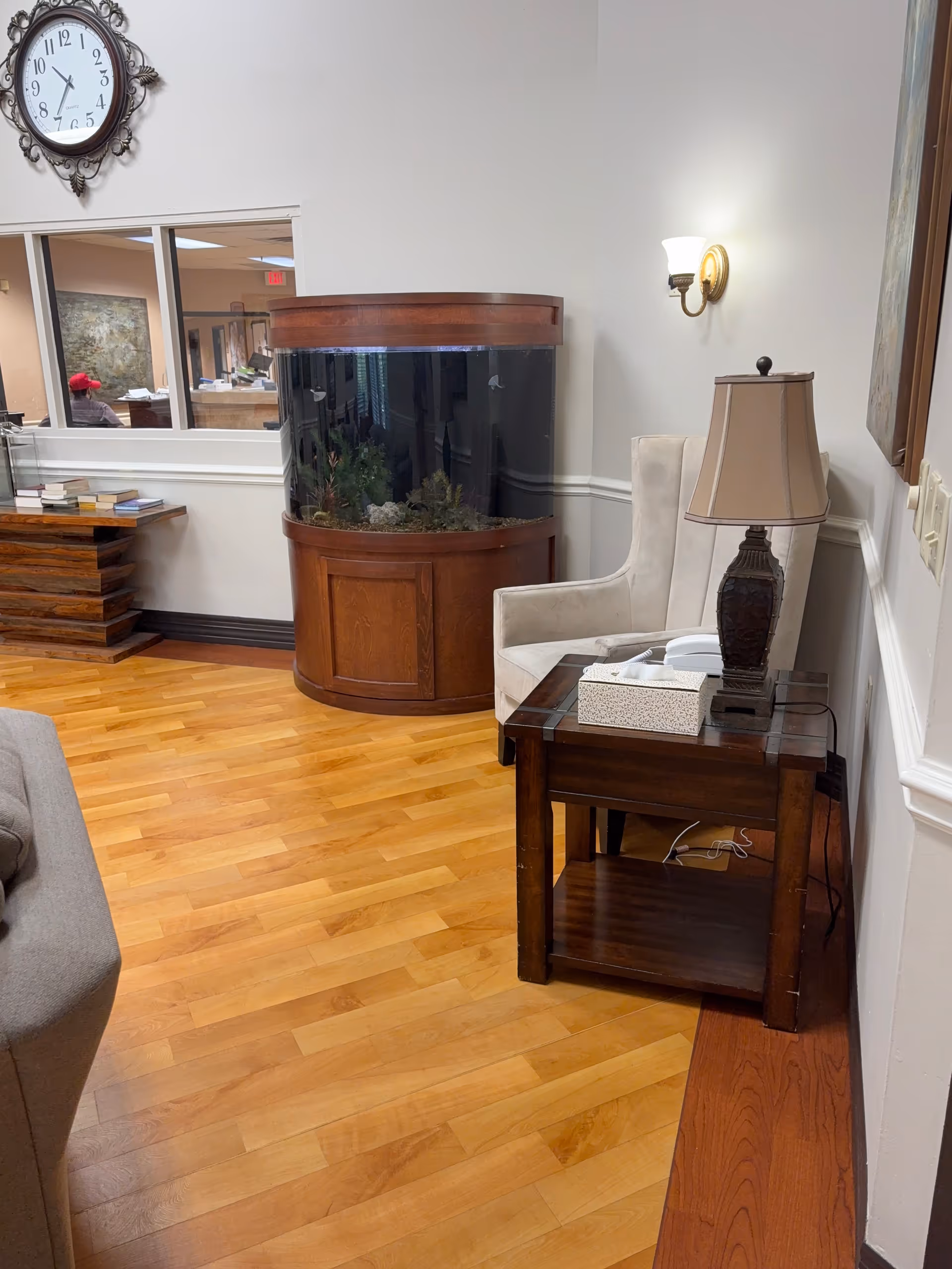 Lobby seating area with a wooden aquarium, armchair, side table and lamp against a wall clock in a senior living facility.