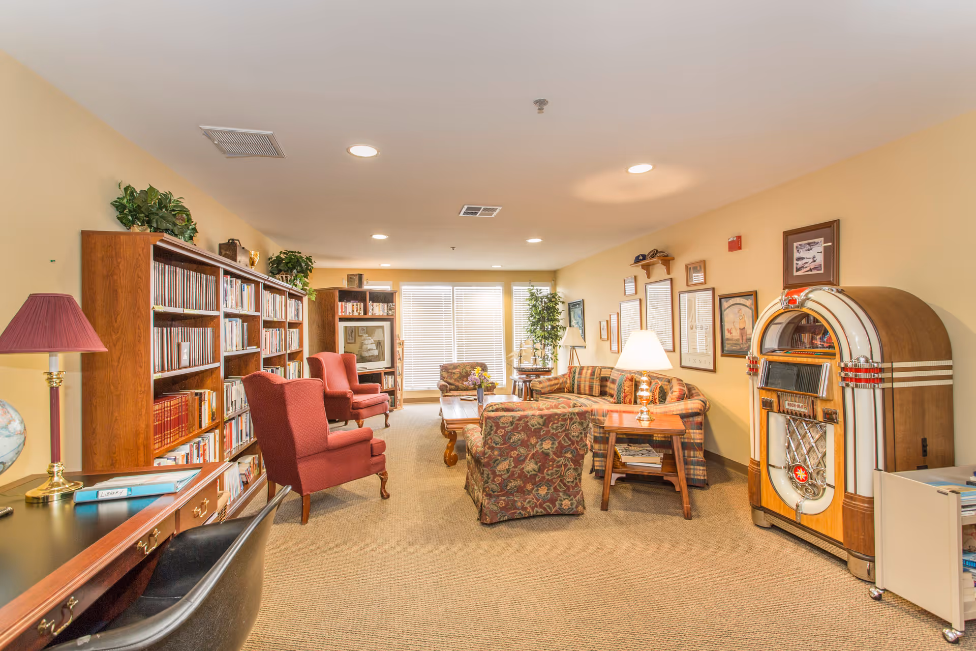A cozy senior living facility common room with bookshelves filled with books on the left, two red armchairs, a floral armchair, a plaid sofa, wooden coffee and side tables, a lamp, framed pictures on the wall, and a vintage jukebox on the right side near a window with blinds letting in natural light.