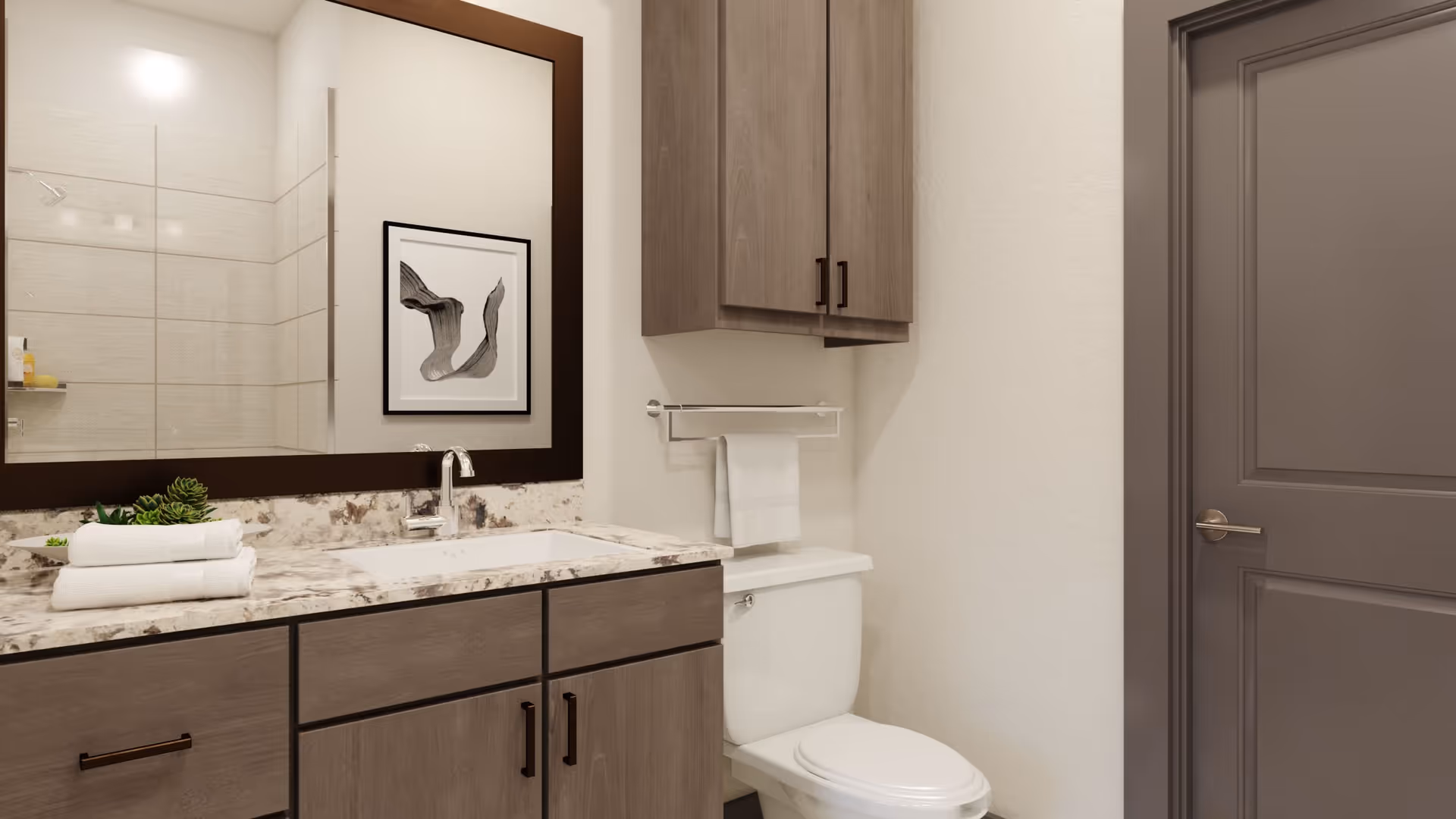 A modern bathroom featuring a white toilet, a wooden cabinet above the toilet, a granite countertop with a sink, a large mirror with a dark frame, and a towel rack with a white towel. There is a framed abstract artwork on the wall reflected in the mirror and a shower area with tiled walls visible in the reflection.