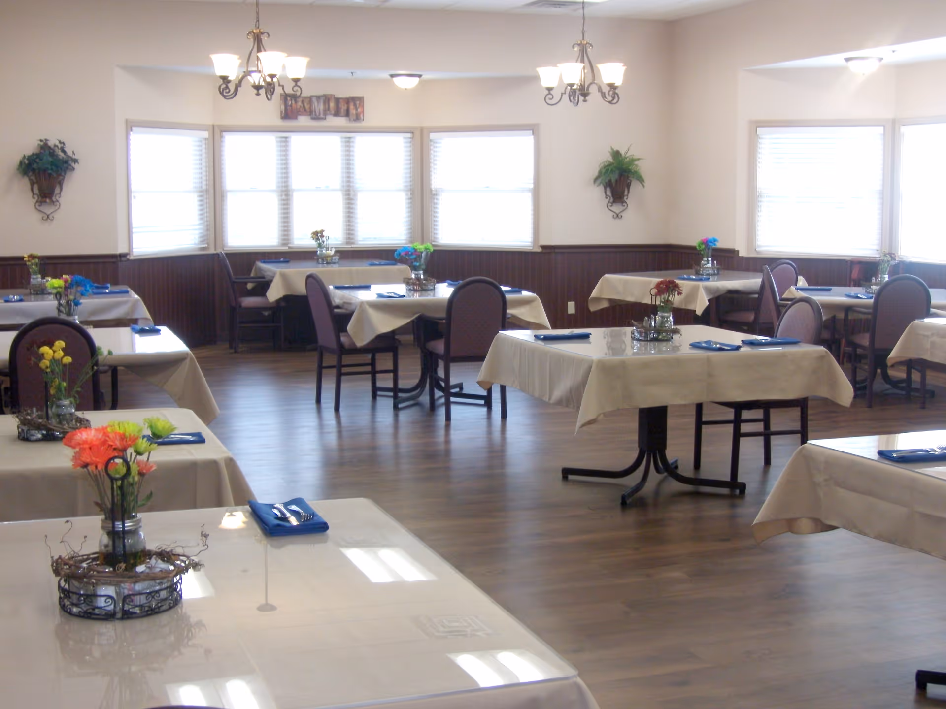 A bright dining room with multiple tables covered in beige tablecloths, each set with blue napkins and silverware. Small flower arrangements in glass jars serve as centerpieces on each table. The room has wooden flooring, large windows with blinds, and decorative wall sconces with plants. Two chandeliers hang from the ceiling providing light.