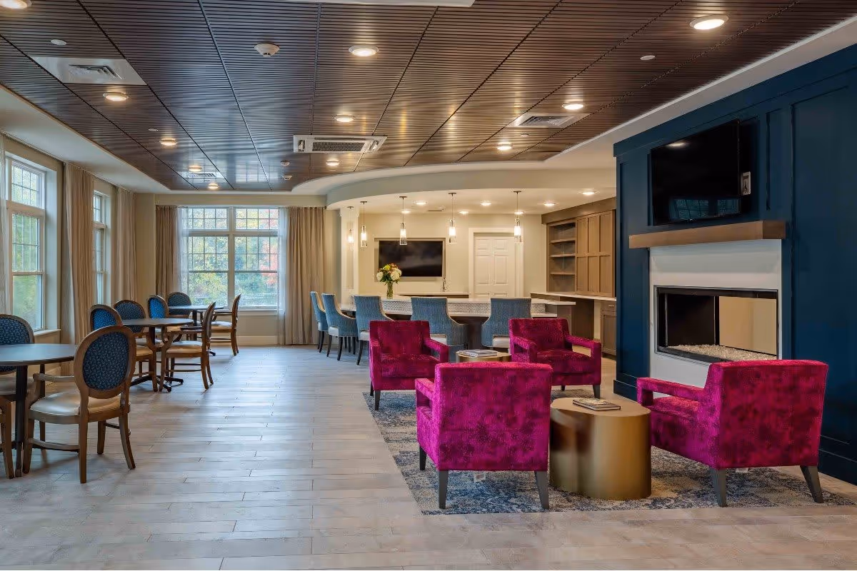 A spacious and modern common area with large windows letting in natural light. The room features a seating area with four bright magenta armchairs arranged around a round gold coffee table on a patterned rug. In the background, there is a bar counter with blue upholstered bar stools and pendant lighting above. A flat-screen TV is mounted above a fireplace set in a dark blue accent wall. Several wooden tables and chairs are positioned near the windows.