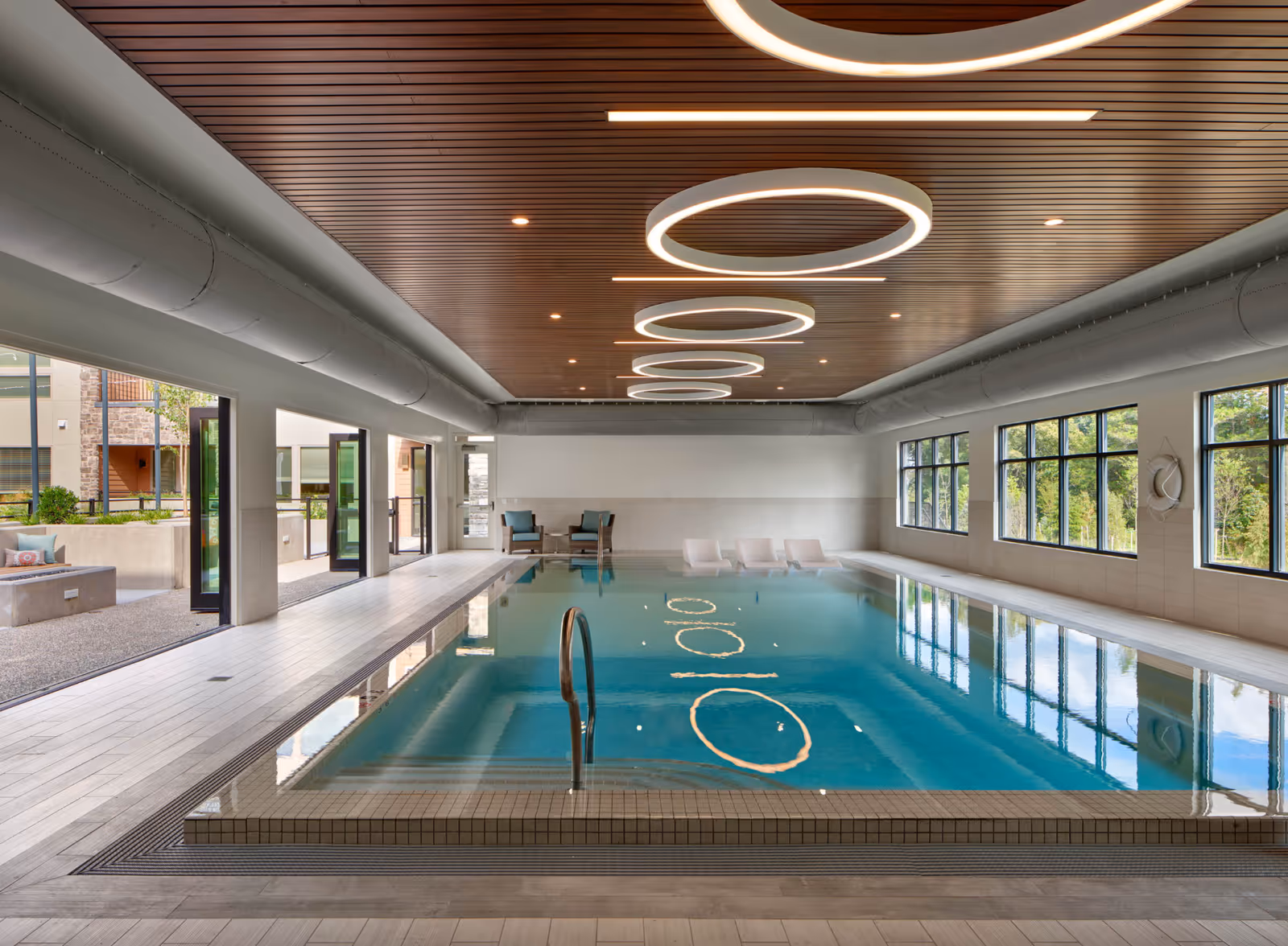 Indoor swimming pool area with modern circular ceiling lights, large windows on the right side, and open doors leading to an outdoor patio area. There are lounge chairs partially submerged in the pool and two armchairs with a small table at the far end of the pool.
