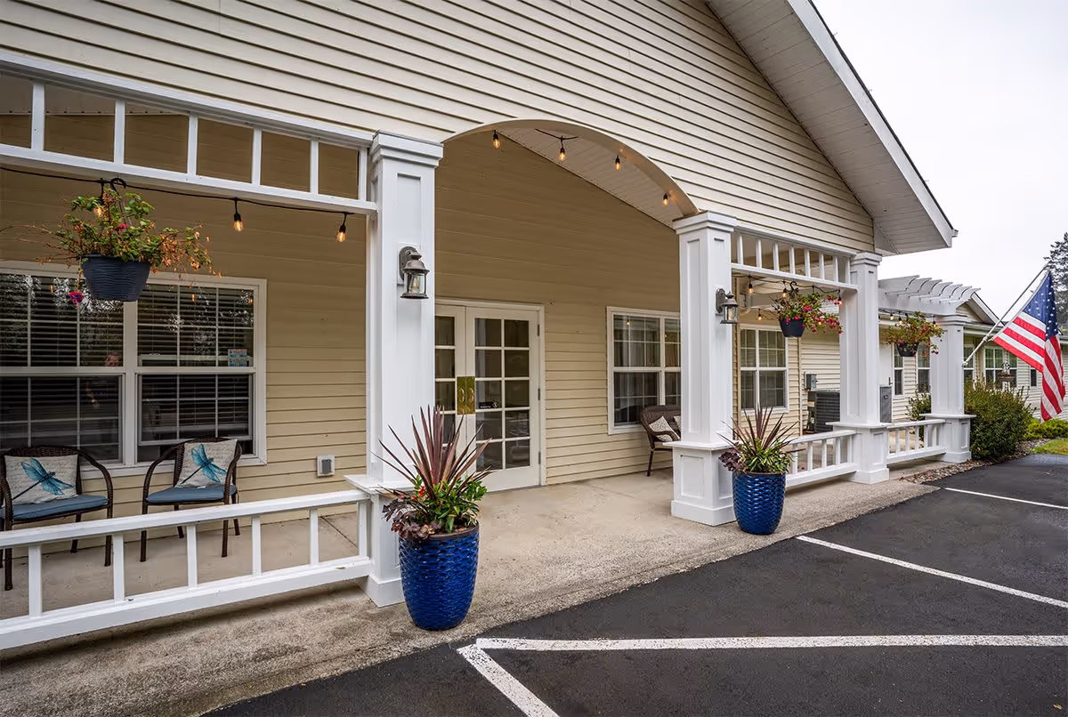 Exterior view of a senior living facility entrance with beige siding, white columns, and a covered porch area. There are hanging flower pots, potted plants in blue containers, chairs with decorative cushions, and an American flag near the parking area.