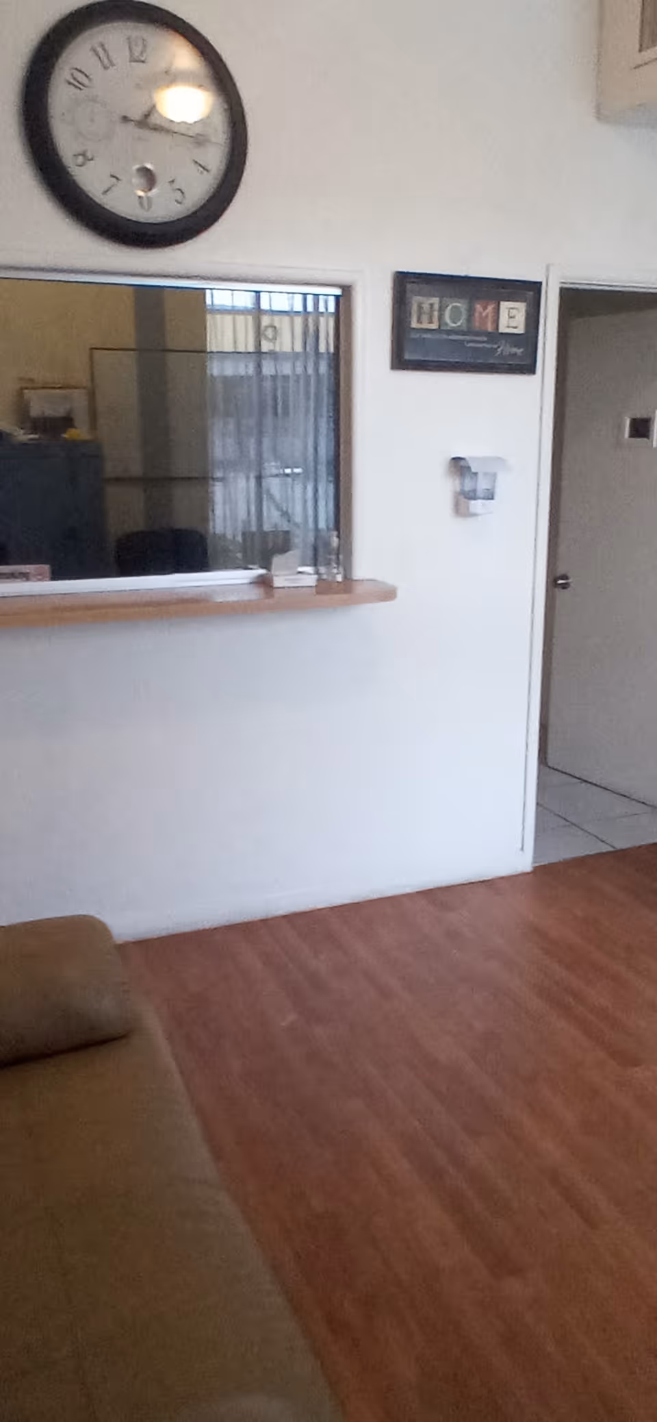 Small reception/living area with a wall clock, pass-through counter, framed 'HOME' sign, and a couch on wood-look flooring.