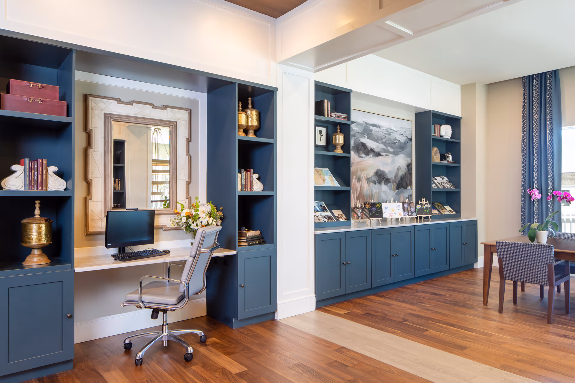 Bright communal living area with built-in blue shelving and cabinets, a desk with a computer and chair, and a small table with chairs and flowers.