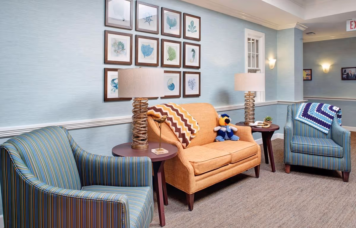 A cozy sitting area in a senior living facility with two striped armchairs and an orange loveseat. Each chair has a crocheted blanket draped over the back. Two side tables with modern spiral-design lamps flank the loveseat. The wall behind features a grid of framed botanical and sea life prints. The room has light blue walls and beige carpeting.