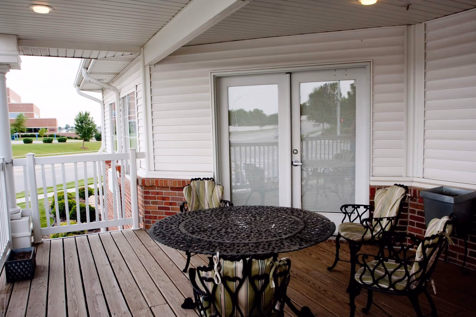 Covered outdoor patio area with a round black metal table and four matching chairs with green striped cushions. The patio has wooden flooring, white railing, and white siding walls with brick accents. Double glass doors lead inside the building, and a grassy area with trees and a building is visible in the background.