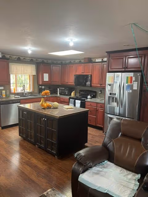 A spacious kitchen with wooden cabinets and a central island. The kitchen features stainless steel appliances including a refrigerator, dishwasher, and microwave. There is a window above the sink with a light curtain, and the floor is wooden. A brown leather recliner chair is partially visible in the foreground with a disposable pad on the seat.