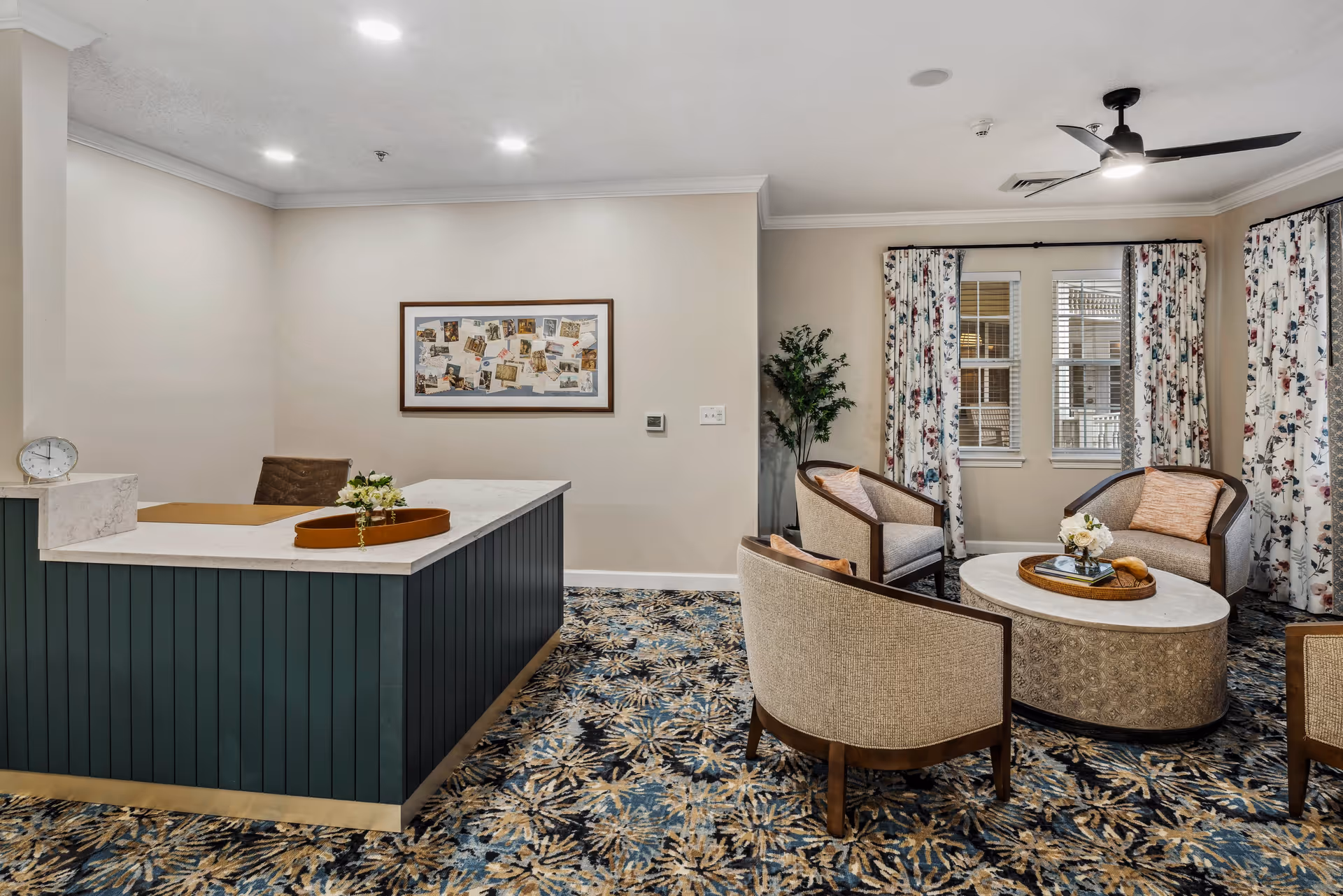 A cozy interior common area with a dark green reception desk on the left and a seating area on the right featuring four beige armchairs around a round coffee table. The room has patterned carpet, floral curtains on the windows, a ceiling fan, and a framed collage on the wall.