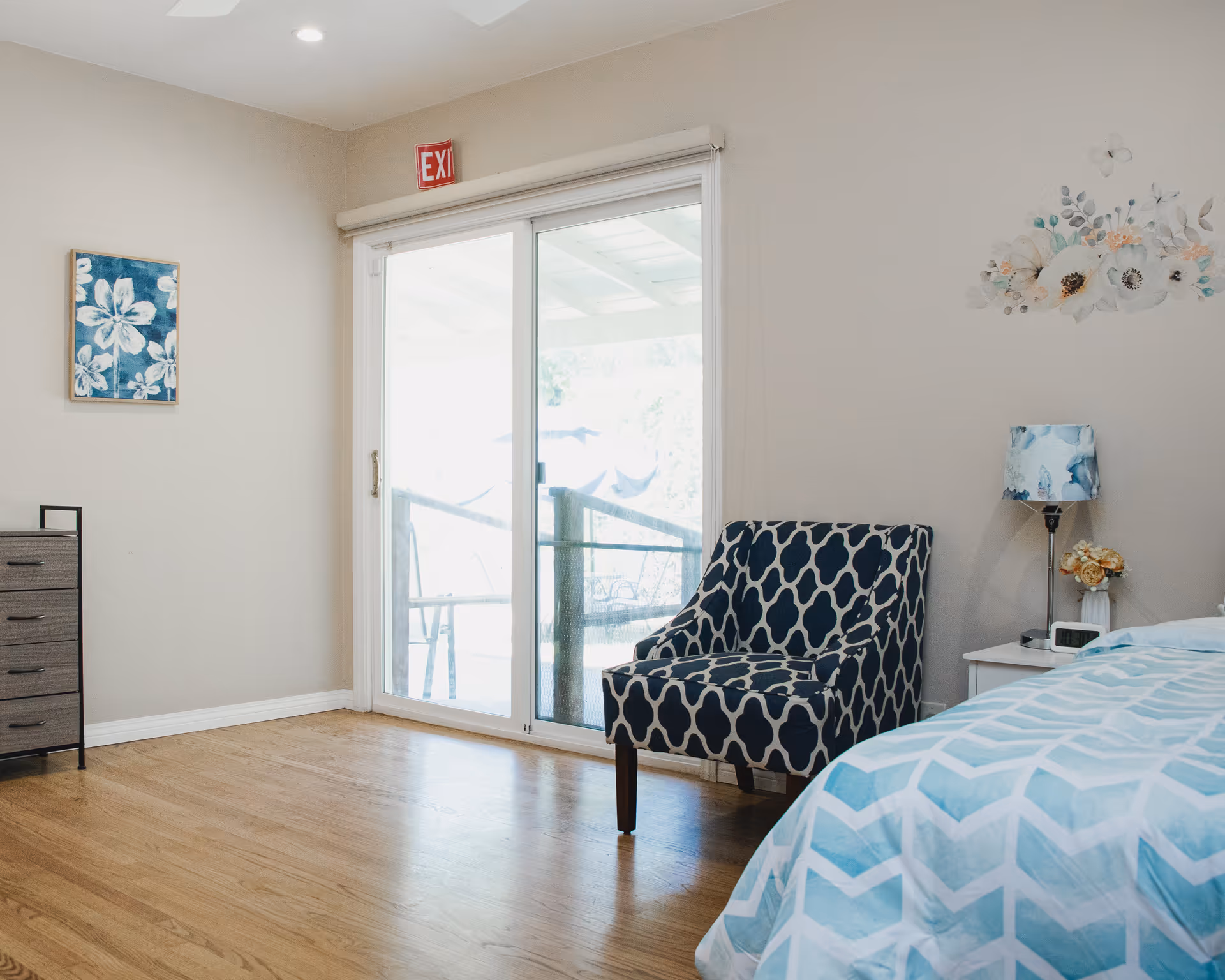 A bright bedroom with wooden flooring featuring a bed with blue and white patterned bedding, a blue and white patterned armchair, a small white nightstand with a lamp and flowers, a chest of drawers, a floral wall painting, and a sliding glass door leading outside with an exit sign above it.