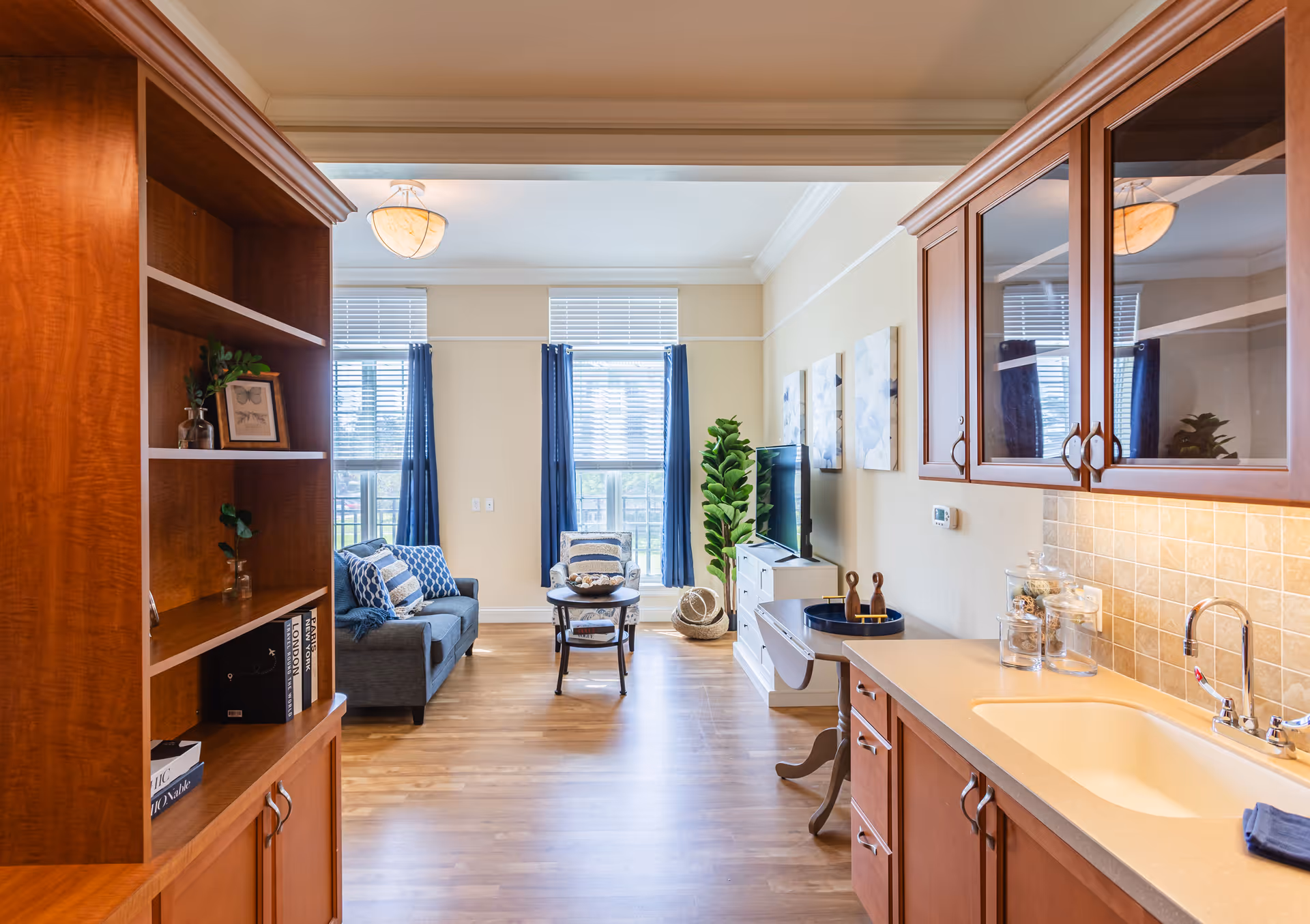 View of a bright living room area from the kitchen, featuring wooden cabinets, a countertop with a sink, a gray sofa with patterned pillows, a small round coffee table, large windows with blue curtains, a TV on a white stand, and decorative plants and artwork on the walls.