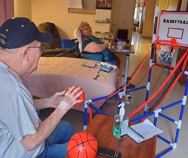 An elderly man sitting and holding a small basketball, preparing to shoot at a mini basketball hoop game set up on a table. An elderly woman is sitting in a wheelchair in the background, watching. The room appears to be a living or common area with a bed, chairs, and a table with hand sanitizer and a notebook.