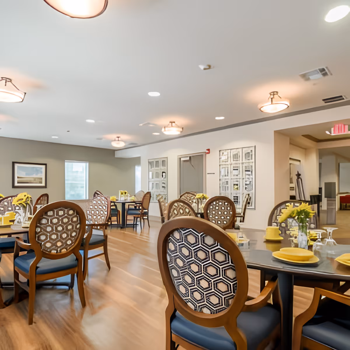 A bright dining room with wooden floors and several round tables set with yellow plates, napkins, and flower centerpieces. The chairs have wooden frames with patterned backs and blue cushions. The room has soft ceiling lights and light-colored walls with windows and framed artwork.