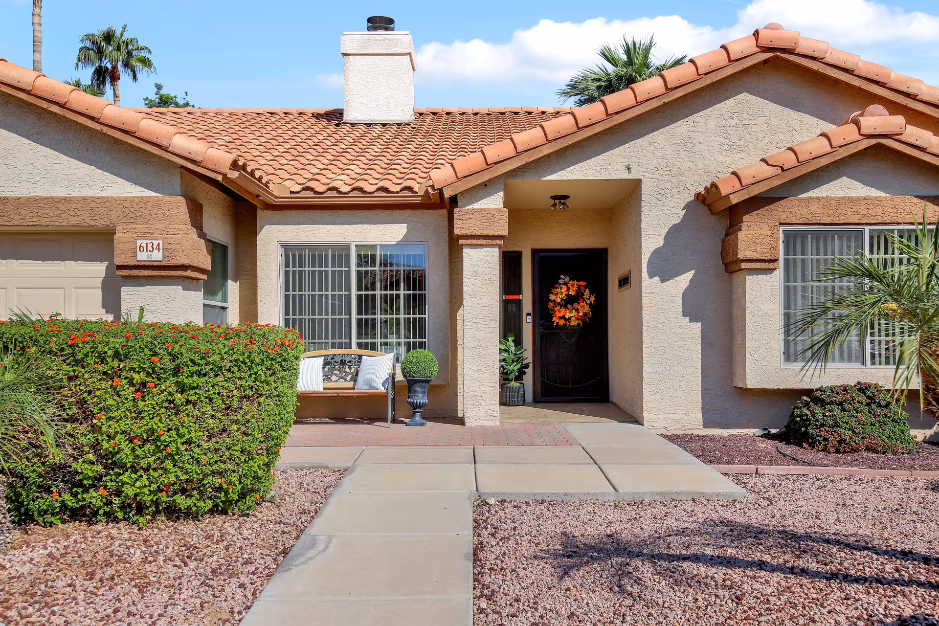 Front exterior view of a single-story house with a tiled roof, beige stucco walls, a black front door decorated with an autumn wreath, large windows with white frames, a small bench with pillows, and landscaped yard with bushes and gravel.