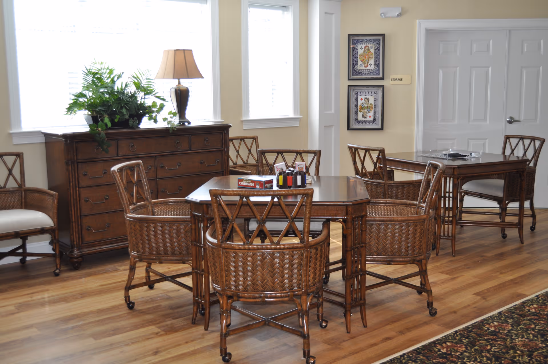 A well-lit room with wooden flooring featuring two wooden tables surrounded by wicker chairs. On one table, there is a Cribbage game box and some card holders. A wooden dresser with a lamp and a potted plant is placed against the wall under two large windows. Two framed pictures hang on the wall next to a door labeled 'Storage'.
