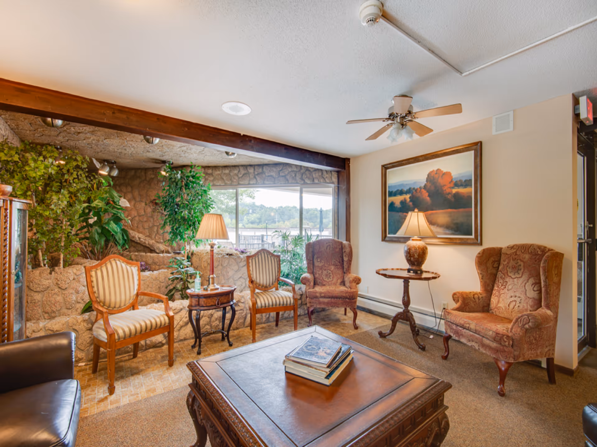 A cozy living room area with a wooden coffee table holding a stack of books in the center. Surrounding the table are two upholstered armchairs with patterned fabric, two wooden chairs with striped cushions, and a black leather chair partially visible. There are two table lamps, one on a small round side table and another on a wooden side table between the striped chairs. The room features a large window with a view of greenery outside, a stone wall with plants, and a ceiling fan with lights. A framed landscape painting hangs on the wall.