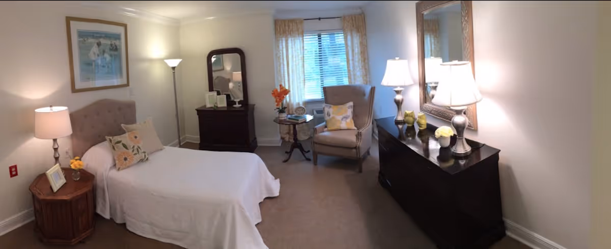 A cozy bedroom in Crowne Place Assisted Living featuring a single bed with floral pillows and a white bedspread. The room includes a wooden nightstand with a lamp and picture frame, a dark wooden dresser with a mirror and two lamps, a comfortable armchair with a floral cushion, a small round table with a flower vase and books, and a window with light-colored curtains letting in natural light.