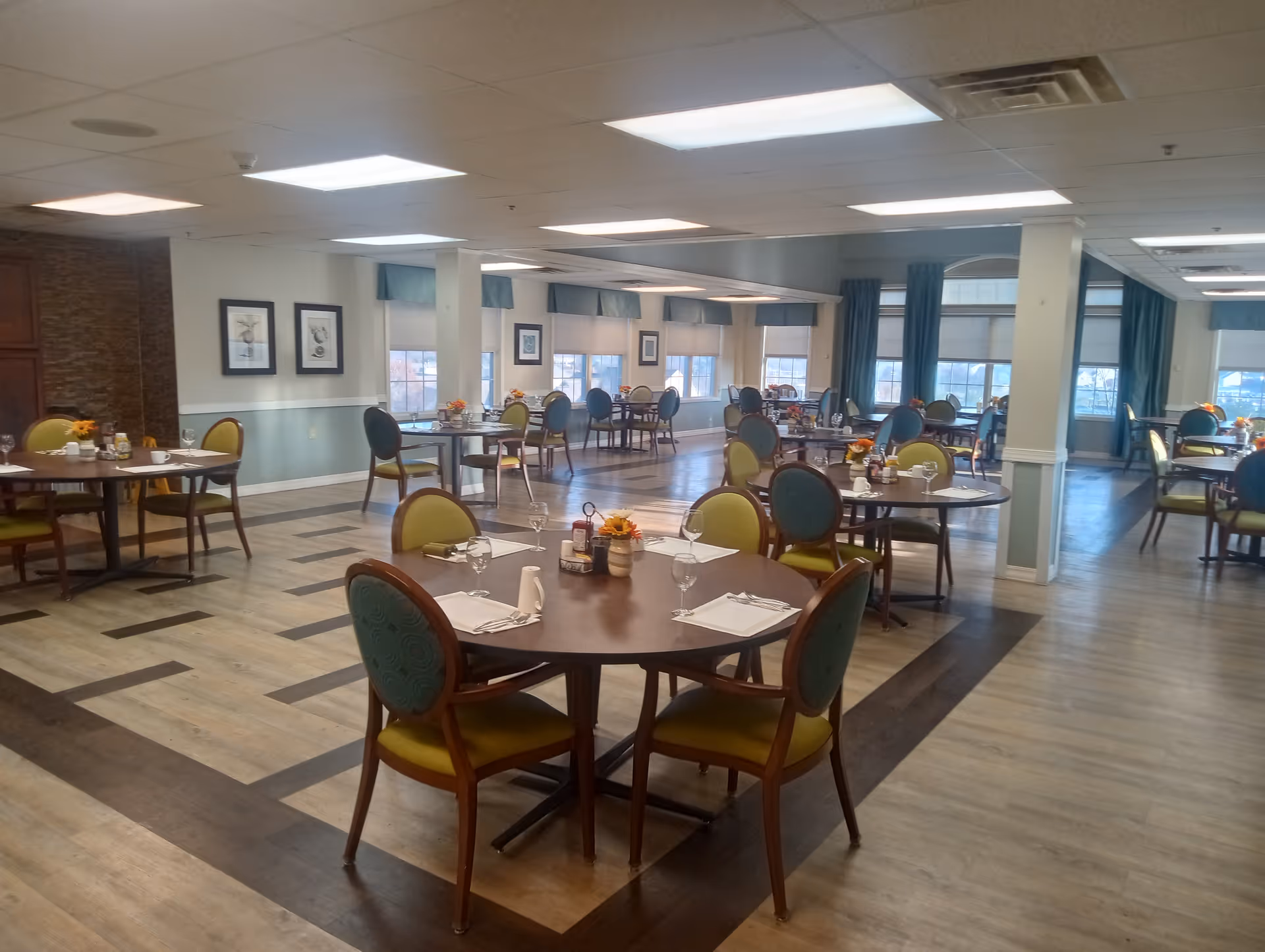 A spacious dining room with multiple round tables set with placemats, napkins, glasses, and small flower centerpieces. The room has large windows with blue curtains, light-colored walls, and wood-patterned flooring with a darker border design. Several chairs with green and blue upholstery surround the tables.