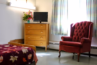 A simple senior bedroom with a red upholstered armchair, a wooden dresser holding a small TV and flowers, and a bed with a floral blanket.