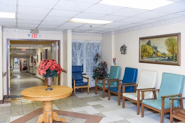 A hallway seating area in a senior living facility with a round wooden table holding a potted plant with red flowers in the foreground. Along the wall are six wooden chairs with cushions in blue, teal, and white colors. The walls are decorated with a framed painting of a landscape and a small wreath. The hallway extends into the background with exit signs visible on the ceiling.
