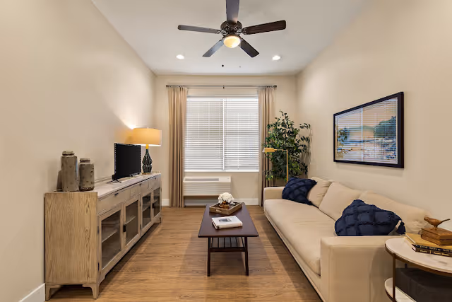 A cozy living room with a beige sofa adorned with two dark blue pillows, a wooden coffee table with decorative items, a wooden TV stand with a small flat-screen TV and a lamp, a ceiling fan with a light, a large window with beige curtains, and a tall green plant in the corner.