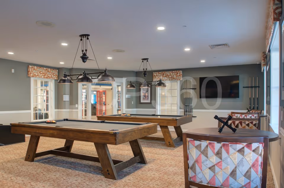 A recreational room with two wooden pool tables, overhead black pendant lights, a wall-mounted TV, and a table with patterned chairs near a window. The room has carpeted flooring and gray walls with white trim.