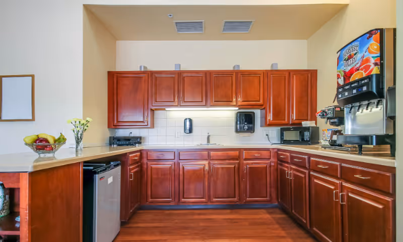 A kitchen area with wooden cabinets and drawers, a countertop with a bowl of fruit and a vase with flowers, a small refrigerator, a microwave, a coffee maker, and a beverage dispenser with colorful fruit graphics.