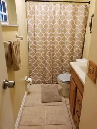 A small bathroom with beige tiled floor, a beige and white patterned shower curtain, a toilet, a wooden vanity with a white countertop and sink, a beige bath mat, and a beige towel hanging on a towel rack. The walls are painted light yellow.