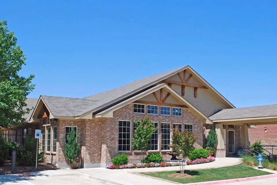 Exterior view of a single-story brick building with large windows, a gabled roof, and landscaped greenery including bushes, small trees, and flowers under a clear blue sky.