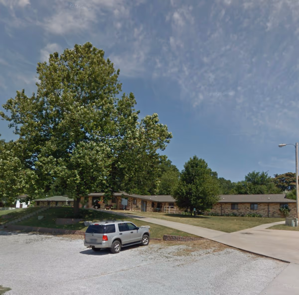 Single-story brick care facility front with a large tree, driveway and a silver SUV parked in a gravel lot.