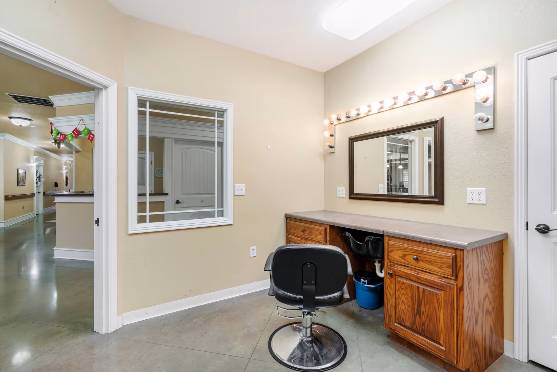 Interior view of a senior living facility room with a wooden vanity desk, a large mirror framed with lights, and a black swivel chair. The room has beige walls and a polished concrete floor. There is a window looking into an adjacent hallway decorated with a festive banner.