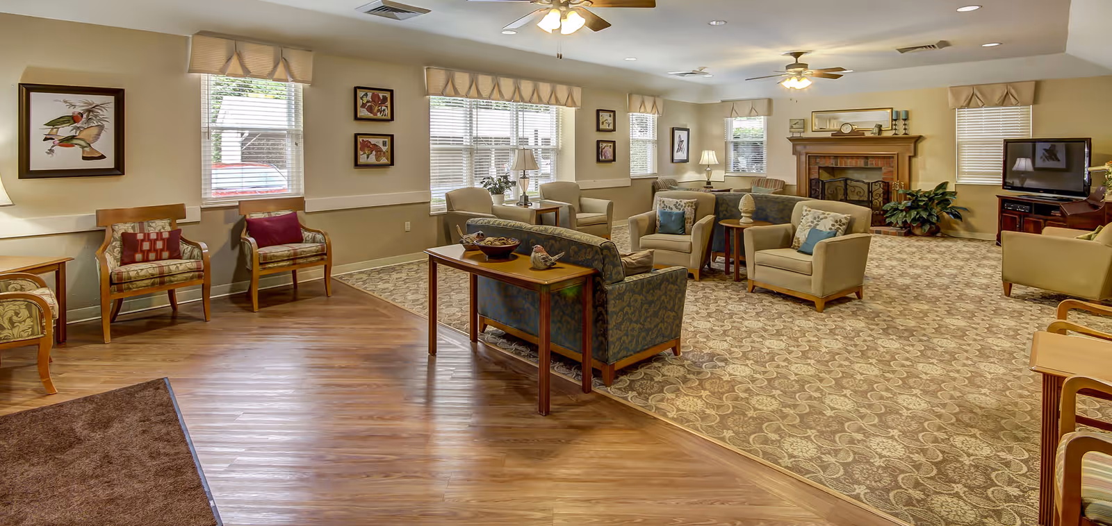 A spacious and well-lit living room area in The Estates of Hidden Lake featuring multiple armchairs and sofas arranged around a wooden coffee table and a fireplace. The room has large windows with blinds, framed artwork on the walls, ceiling fans, and a television on a stand. The flooring is a combination of wood and patterned carpet.
