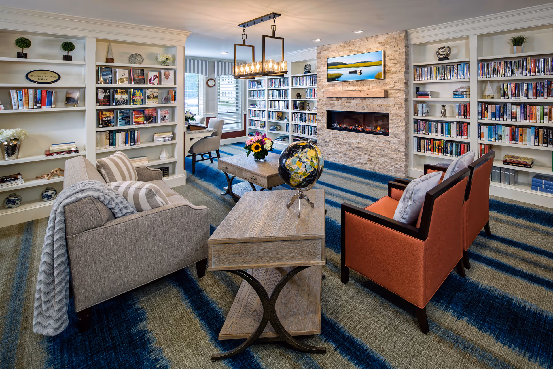 A cozy communal living room/library with bookshelves, a stone fireplace, seating arrangements, and a central coffee table with a globe.