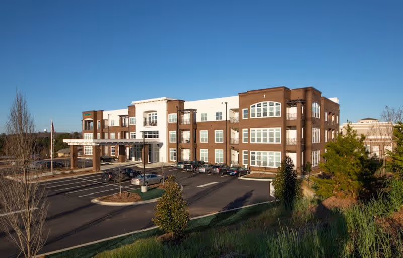 Exterior view of a three-story senior living facility building with a parking lot in front, surrounded by landscaped greenery and trees under a clear blue sky.
