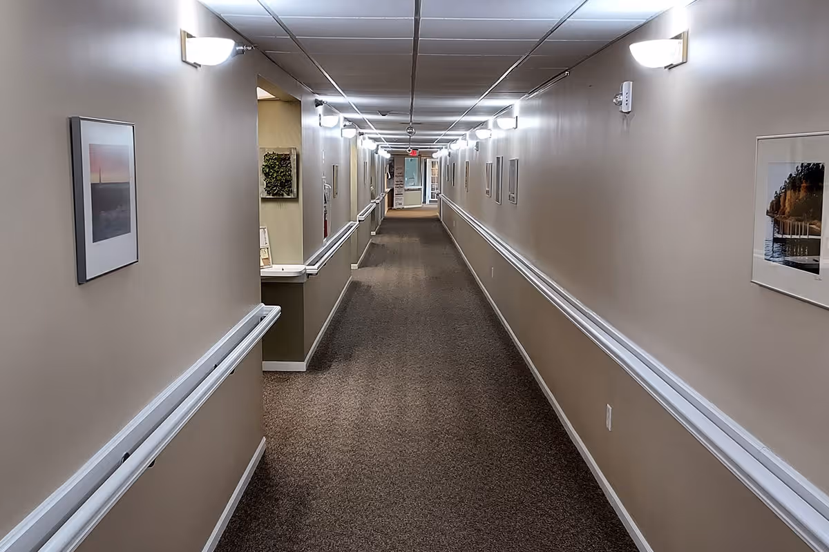 Long carpeted hallway with handrails, framed artwork, and wall-mounted lights in a senior living facility.
