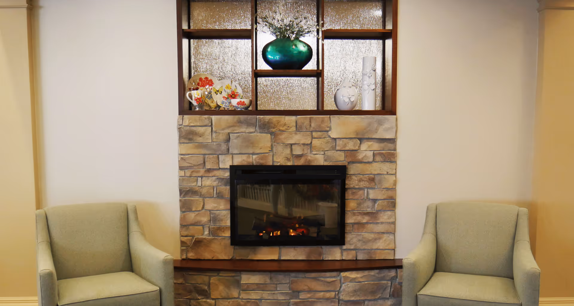 A cozy seating area with two light green armchairs placed on either side of a stone fireplace. Above the fireplace is a wooden shelf with decorative items including a green vase with flowers, floral plates, and white ceramic vases. The walls are painted a soft beige color.