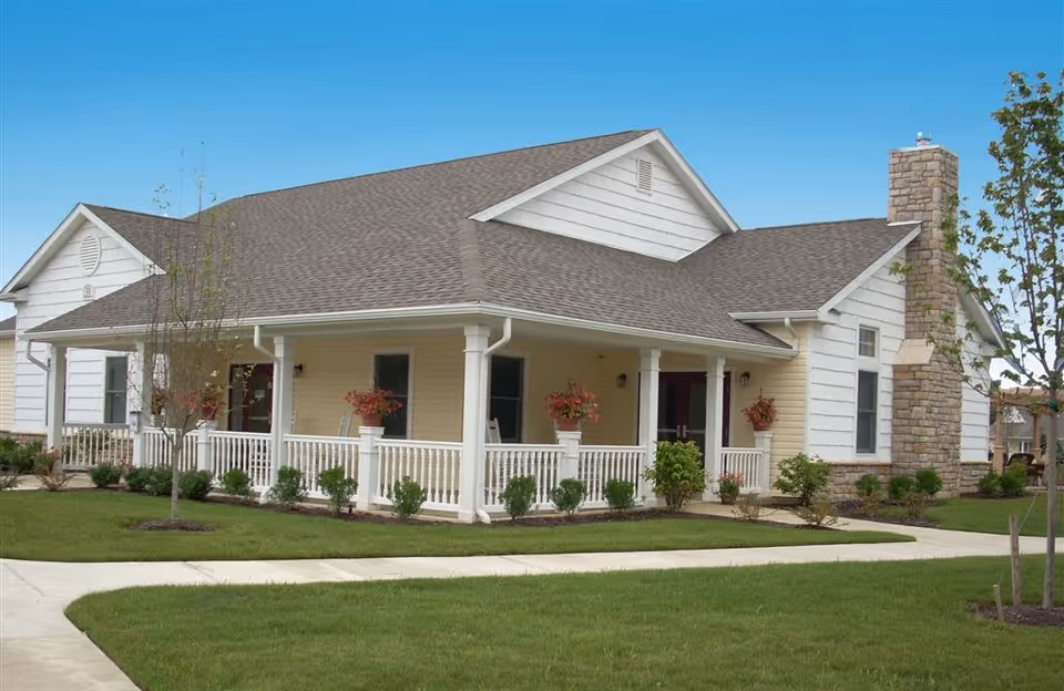 Single-story light-colored building with a covered wraparound porch, white railings, a stone chimney, and a manicured lawn and walkway.