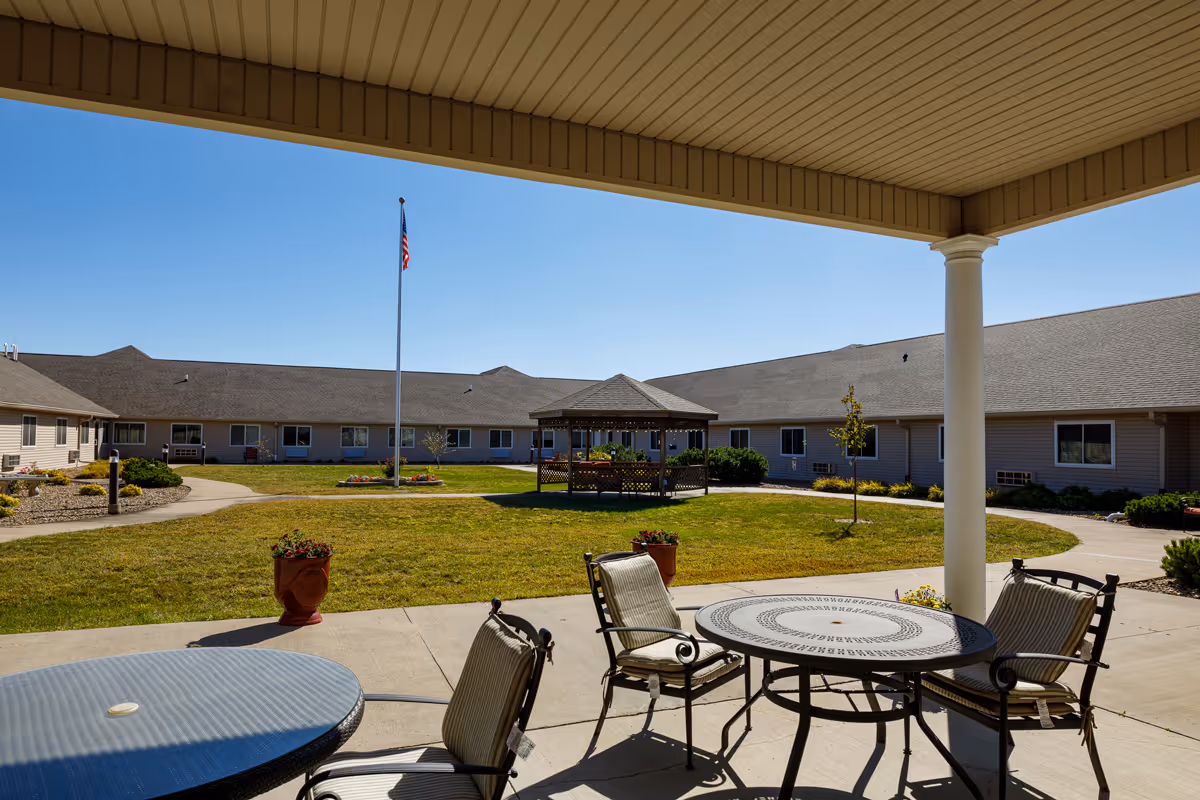 Outdoor courtyard area of Homestead Assisted Living & Memory Care of Oskaloosa featuring a covered patio with round tables and cushioned chairs. The courtyard has a grassy lawn, a gazebo in the center, a flagpole with an American flag, and a clear blue sky overhead. The building surrounds the courtyard with multiple windows visible.