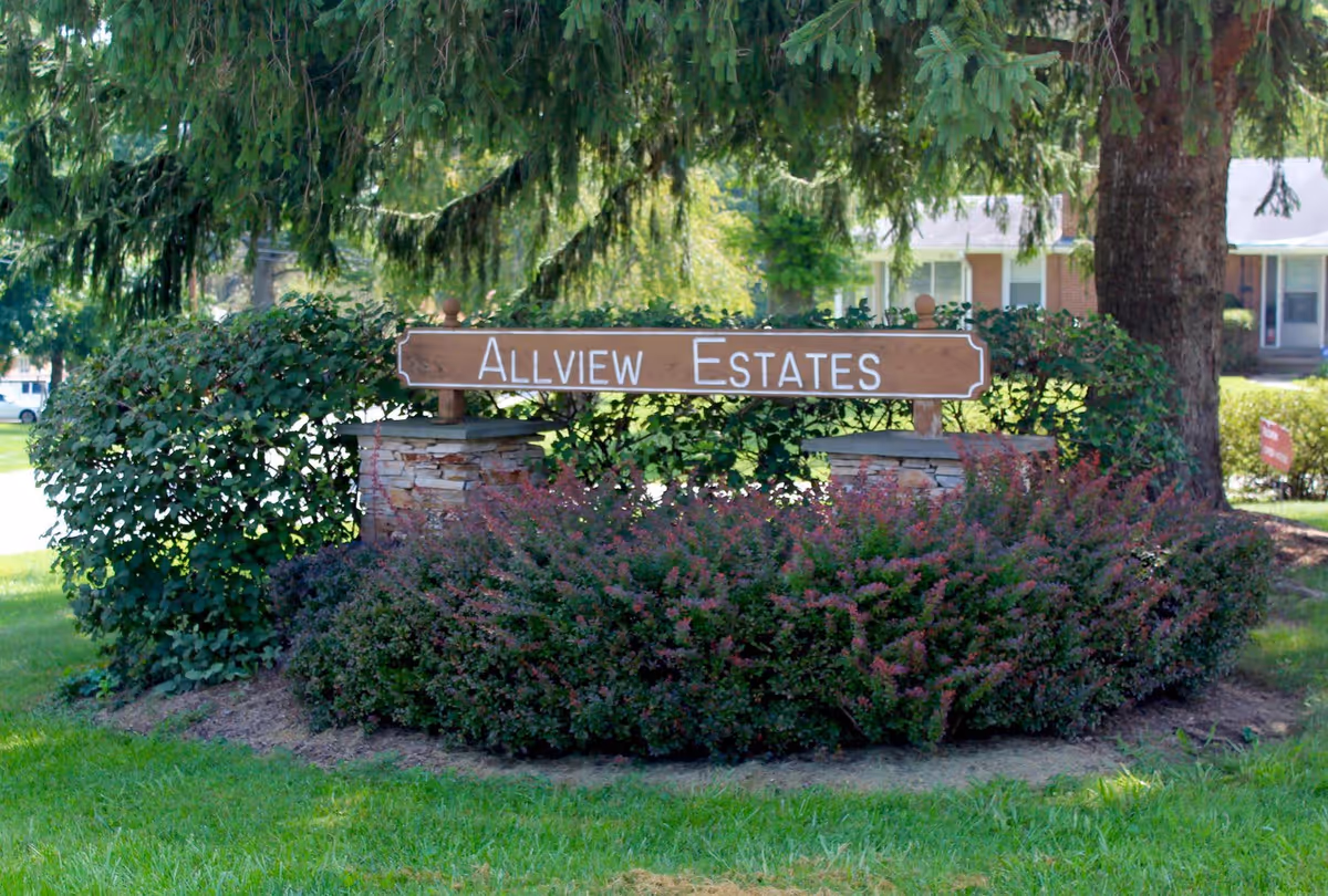 A wooden sign reading 'Allview Estates' mounted on two stone pillars surrounded by green bushes and plants, with a large tree and residential buildings in the background.