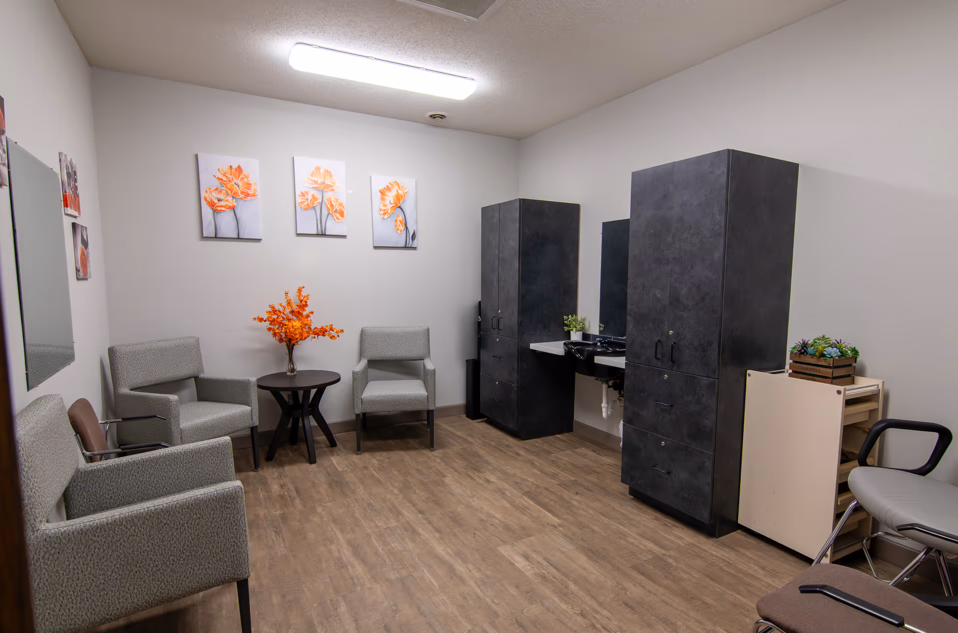 A small room with four gray upholstered chairs arranged around a small round table with an orange flower arrangement. The walls are white with three orange flower paintings. There are two tall dark cabinets and a small sink with a mirror on the right side. The floor is wood laminate.
