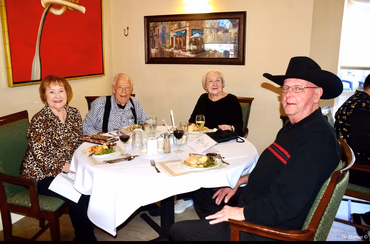 Four elderly people sitting around a dining table with white tablecloth, enjoying a meal together in a well-lit room with artwork on the walls. The group includes three women and one man wearing a black cowboy hat and glasses. Plates with food and glasses of wine and water are on the table.