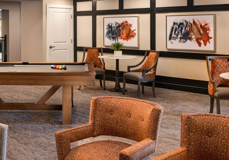 A cozy indoor lounge area featuring a wooden pool table with billiard balls set up, surrounded by several upholstered chairs in a warm orange pattern. There are small round tables with potted plants and abstract artwork hanging on the walls. The room has a carpeted floor and neutral-colored walls with dark trim.