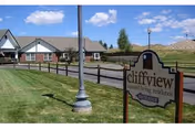 Exterior view of a Cliffview Assisted Living Center sign in front of a single-story brick building with lawn, fence, and blue sky.