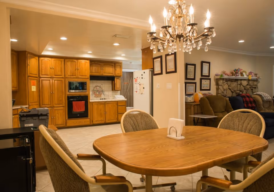 Interior view of a senior living facility showing a wooden dining table with four cushioned chairs around it. In the background, there is a kitchen area with wooden cabinets, a built-in oven, a microwave, and a refrigerator. To the right, there is a living room area with armchairs, a stone fireplace decorated with flowers, and framed certificates on the wall. A chandelier hangs from the ceiling above the dining table.