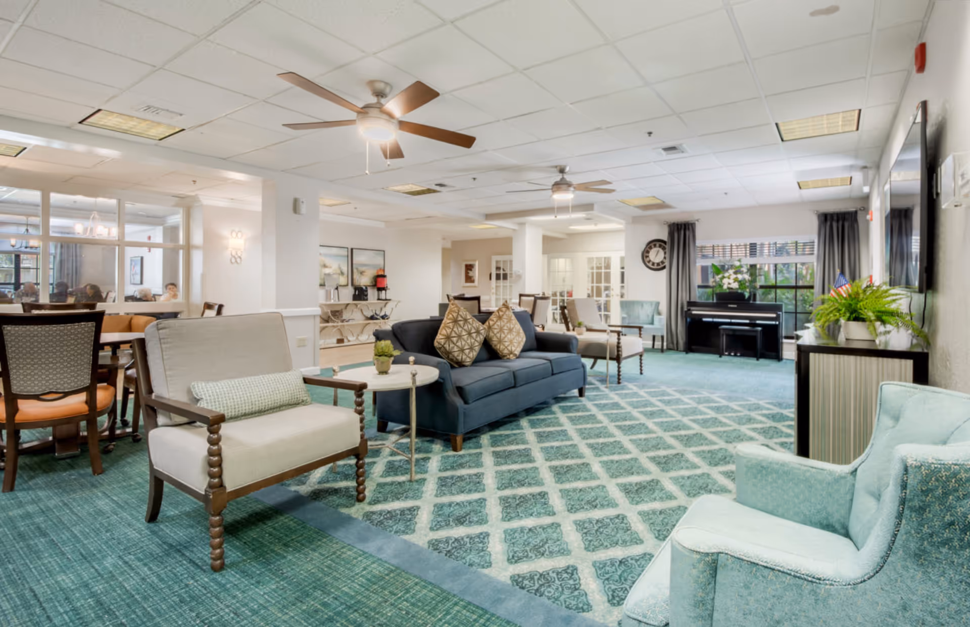 Spacious senior living common room with sofas and armchairs arranged on a patterned carpet, a piano near the window, dining tables, and ceiling fans.